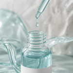 A drop of blue copper peptide serum falling from the glass dropper into the open 30 mL bottle