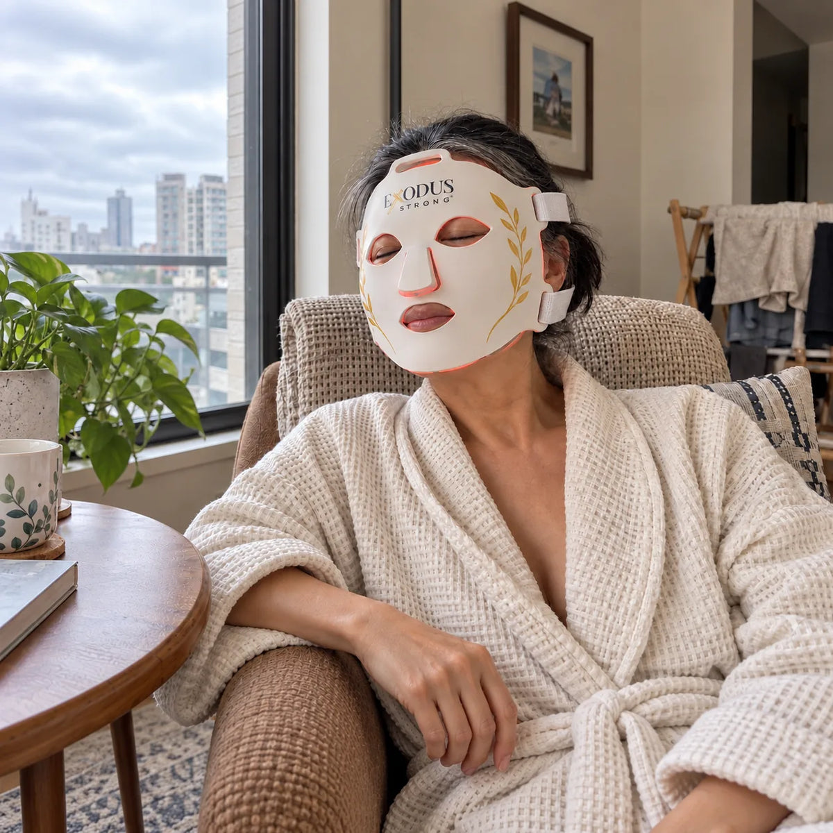 A woman in a robe by a city window wearing the white Red Light+ Face Mask