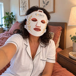 A woman sitting on her bed wearing the white Red Light+ Face Mask