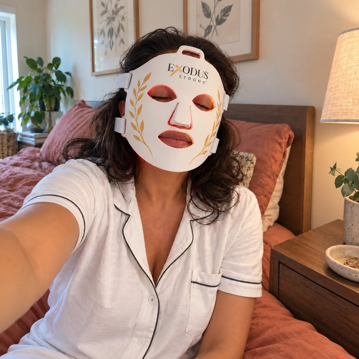 A woman sitting on her bed wearing the white Red Light+ Face Mask