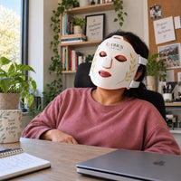 A woman taking a break at her desk wearing the white Red Light+ Face Mask
