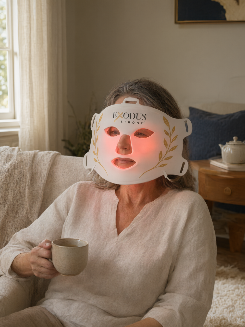 A woman in a reading chair wearing the Red Light+ Face Mask with tea