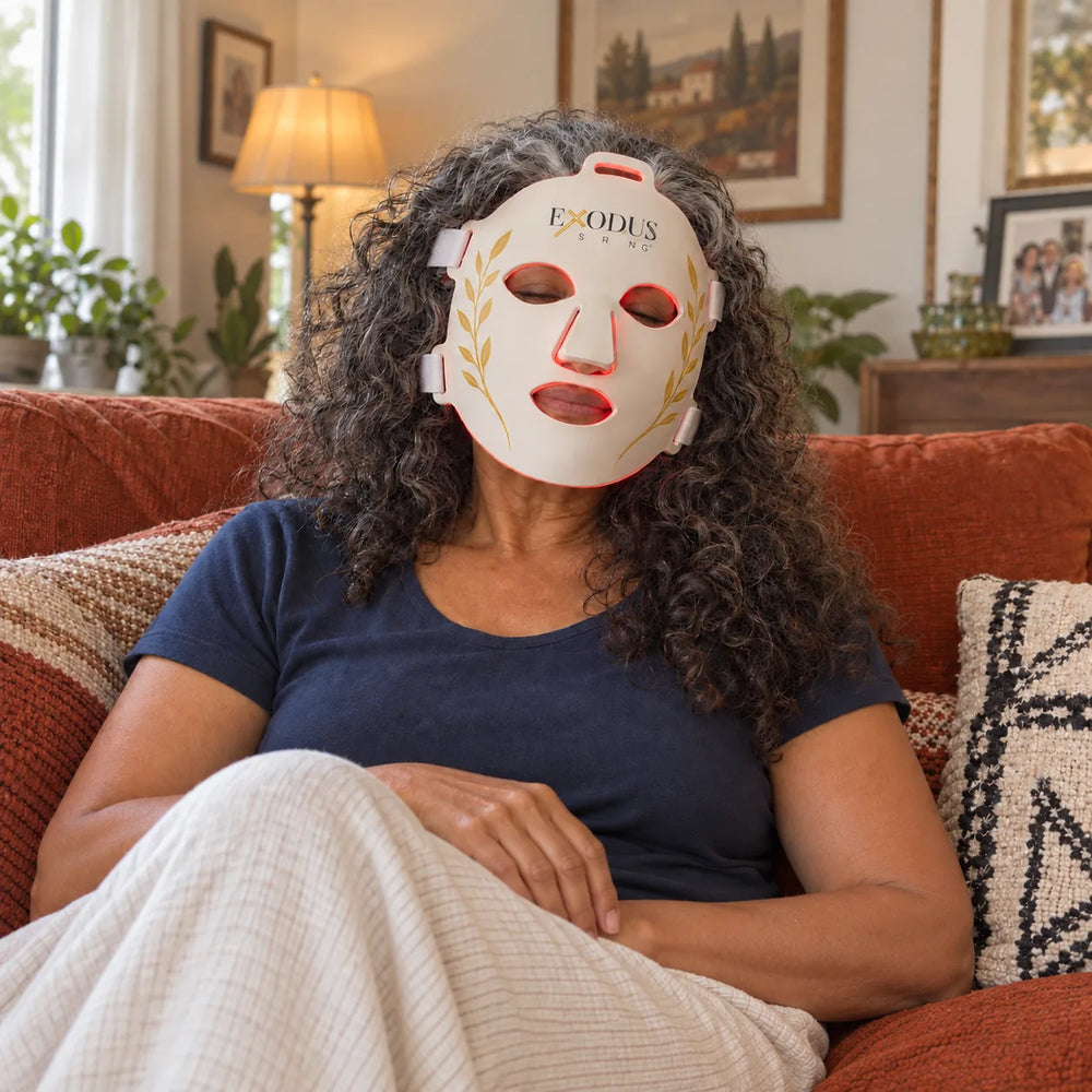 A woman with curly grey hair on her sofa under a blanket wearing the white Red Light+ Face Mask